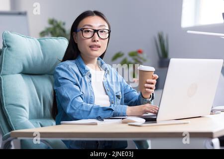 Kaffeepause im Büro. Junge, schöne asiatische Geschäftsfrau, die in einem modernen Büro eine Tasse Kaffee in der Hand hält und Kaffee trinkt. Er schaut lächelnd auf die Kamera. Stockfoto
