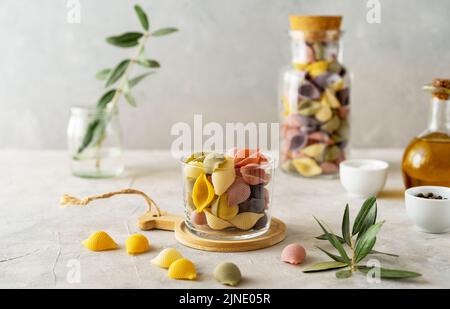 Mehrfarbige italienische Pasta Conchiglie oder Muscheln in Glas und Flasche, Olivenöl, Salz, Pfeffer, Olivenzweig auf Betongrund Stockfoto