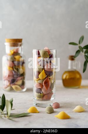 Mehrfarbige italienische Pasta Conchiglie oder Muscheln in Glas und Flasche, Olivenöl, Olivenzweig. Konkreter Hintergrund Stockfoto
