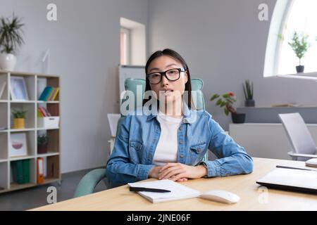 Fernstudium. Eine junge Asiatin ist Lehrerin, führt Online-Kurse, Unterricht durch. Er schaut in die Kamera, erklärt, erzählt. Sitzen in Brille und Jeans an einem Tisch an einem Computer in einem Klassenzimmer Stockfoto