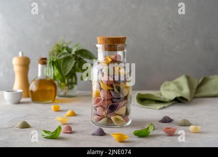 Mehrfarbige italienische Pasta Conchiglie oder Muscheln in Glasflasche, Olivenöl, Olivenzweig. Konkreter Hintergrund Stockfoto