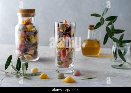 Mehrfarbige italienische Pasta Conchiglie oder Muscheln in Glas und Flasche, Olivenöl, Olivenzweige. Konkreter Hintergrund Stockfoto