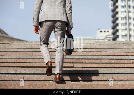 Mann in einem Business-Anzug mit Ledertasche, der in der Stadt Steintreppen klettert, männliche Beine in Bewegung auf den Stufen. Konzept von Karriere, Erfolg Stockfoto