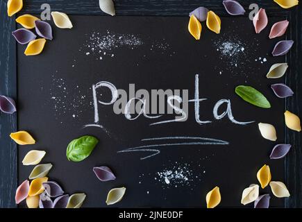 Mehrfarbige italienische Pasta-Conchiglie oder Muscheln mit Basilikumblättern auf Kreidetafel. Wort Pasta schrieb mit Kreide Stockfoto