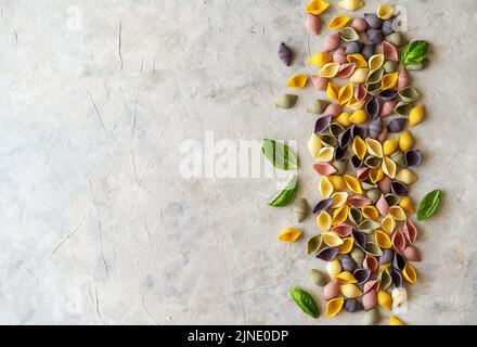 Mehrfarbige italienische Pasta-Conchiglie oder Muscheln mit Basilikumblättern auf betontem Hintergrund. Flach mit Kopierplatz. Stockfoto