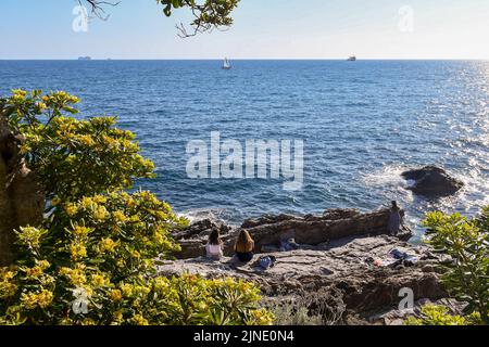 Meeresblick von der Promenade mit Mädchen, die auf den Felsen am Meer sitzen und blühenden Pflanzen im Vordergrund, Nervi, Genua, Ligurien Stockfoto