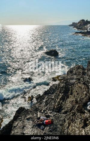 Blick auf den Golf des Paradieses mit einem Mann, der auf den Felsen am Meer liegt, und dem Fischerdorf im Hintergrund, Nervi, Genua, Ligurien, Italien Stockfoto