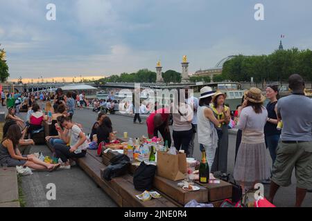 Menschen, hauptsächlich Touristen, treffen sich mit Essen und Wein entlang der seine am Pont Alexandre III. Schöner Sommertag im Freien in Paris, Frankreich. Stockfoto