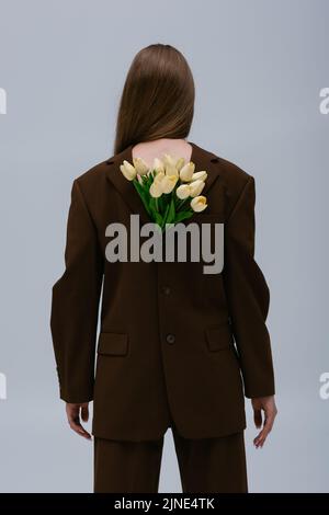 Rückansicht des Teenage-Models in braunem Blazer mit Tulpen isoliert auf grau Stockfoto