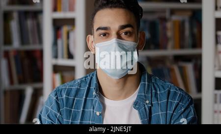 Nahaufnahme ernsthaft nachdenklichen jungen Mann allein im Haus sitzen mit schützenden OP-Maske hispanic guy Student Blick auf Kamera posiert in medizinischen Stockfoto
