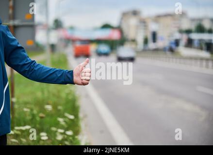 Nahaufnahme der Menschenhand, die am Straßenrand anhält. Männliche Hand zeigt Daumen nach oben Geste im Freien auf unscharfem Hintergrund. Anhalter, Anhalter, Auto-Stopp-Konzept. Stockfoto