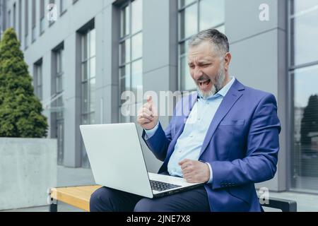 Erfolgreicher grauhaariger Geschäftsmann, der am Laptop arbeitet und den Siegestriumpf feiert, übergewichtiger Mann, der vor dem Bürogebäude auf der Bank sitzt Stockfoto