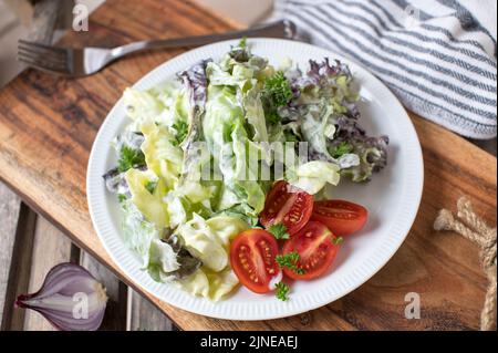 Beilagensalat mit Salat und Tomaten auf einem mit Sauerrahm-Dressing marinierten Teller. Traditionelle deutsche Beilage oder Vorspeise Stockfoto