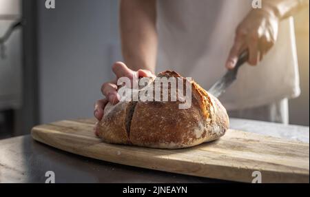 Ein männlicher Bäcker schneidet frisch gebackenes Brot mit einem Messer, um die Qualität zu überprüfen. Herstellung von Backwaren als kleines Unternehmen. Stockfoto
