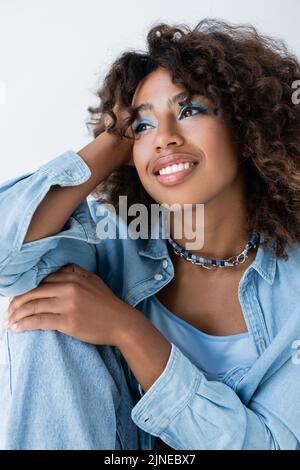 Fröhliche afroamerikanische Frau in Perlen und blauem Denim-Hemd, die isoliert auf Grau davonschaut Stockfoto