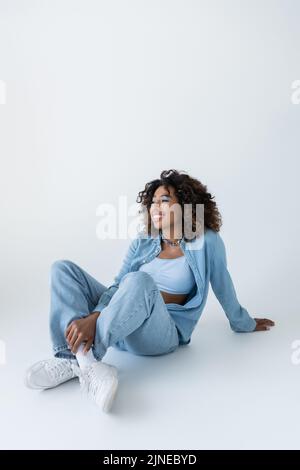 Glückliche afroamerikanische Frau in Jeans und Sneakers, die mit gekreuzten Beinen auf grauem Hintergrund sitzt Stockfoto