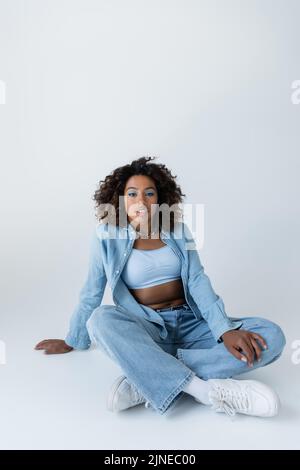Brünette afroamerikanische Frau in blauen Denim-Kleidung Blick auf die Kamera, während sie auf grauem Hintergrund sitzt Stockfoto