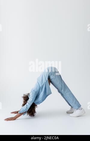 Seitenansicht einer afroamerikanischen Frau in Denim-Kleidung, die in einer Delphin-Pose auf grauem Hintergrund steht Stockfoto