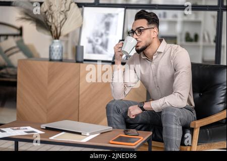 Mann, der Kaffee trinkt, sitzt zu Hause auf einem Stuhl Stockfoto