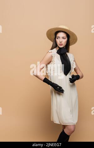 Trendige junge Frau in Vintage-Kleid und Sonnenhut posiert isoliert auf Beige Stockfoto