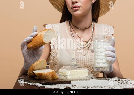 Beschnittene Ansicht einer jungen Frau in Handschuhen, die Brot und Milch in der Nähe von Butter hält, die auf Beige isoliert ist Stockfoto