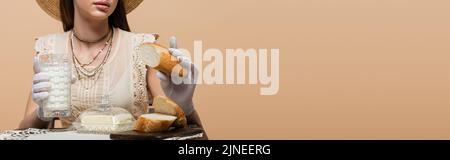 Beschnittene Ansicht einer Frau in Handschuhen, die Milch und Brot in der Nähe von Butter hält, isoliert auf beigem Banner Stockfoto