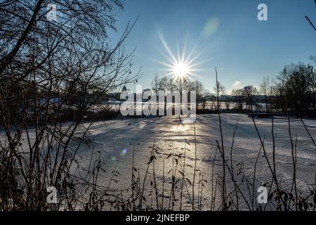 Die Sonne scheint hell über einem gefrorenen See, gesehen durch trockene Pflanzen und Äste Stockfoto