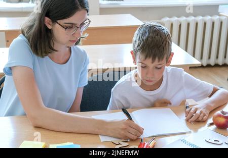 Ein Junge und eine Lehrerin sitzen an einem Tisch mit Heften und Stiften. Eine Mutter hilft ihrem Sohn, in ein Notizbuch zu schreiben. Der Babysitter hilft, schoo zu vervollständigen Stockfoto