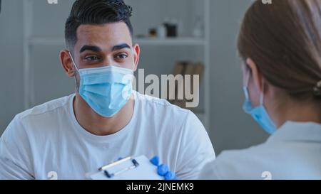 Arabischer Mann hispanic Kerl Patient krank männlich trägt medizinische Schutzmaske auf Gesicht spricht über Symptome von gesundheitlichen Problemen zu unkenntlich Arzt Frau Stockfoto