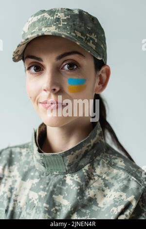 Militärfrau mit ukrainischer Flagge auf Gesicht gemalt Blick auf Kamera isoliert auf grau, Stock Bild Stockfoto