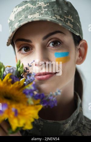 Porträt einer positiven Militärfrau, die die Kamera in der Nähe von Blumen auf grauem, stockem Bild betrachtet Stockfoto