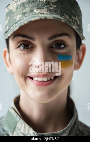 Nahaufnahme Porträt einer Militärfrau mit ukrainischer Flagge auf dem Gesicht lächelnd auf die Kamera isoliert auf grauem, Stock-Bild Stockfoto