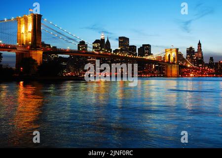 Die Beleuchtung der Brooklyn Bridge spiegelt sich im Wasser des East River wider, wenn sich die Dämmerung auf der Skyline von Manhattan nähert Stockfoto