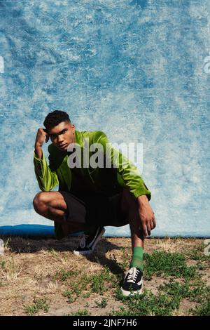 In voller Länge afroamerikanischer Mann in grünem Blazer und Sneakers posiert auf Haunches in der Nähe von blauen Vorhapern, Stockbild Stockfoto