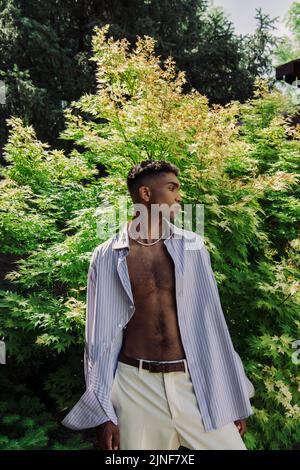 Junger afroamerikanischer Mann im gestreiften Hemd, der in der Nähe des grünen Buschs im Park steht, Stockbild Stockfoto