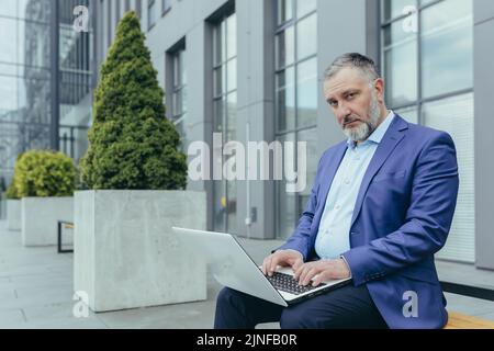 Porträt eines älteren grauhaarigen Geschäftsmannes vor dem Bürogebäude, eines Mannes, der am Laptop arbeitet und tippt, der nachdenklich und ernsthaft auf die Kamera schaut Stockfoto