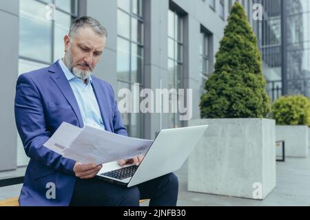 Seriöser, grauhaariger Geschäftsmann, Investor mit Papierkram, der vor dem Bürogebäude auf der Bank sitzt, Mann im Geschäftsanzug mit Dokumenten und Laptop, der arbeitet Stockfoto