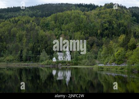 Wunderschöne Landschaften in Norwegen. Weste-Agder. Wunderschöne Landschaft der White Gyland Kirche, die sich im See spiegelt. Berge, Straße und Bäume im Hintergrund Stockfoto