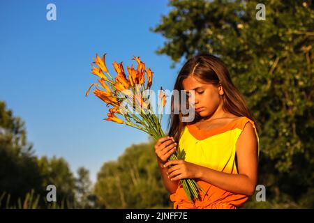 Ein schönes nachdenkliches Teenager-Mädchen in gelbem Kleid mit einem Strauß oranger Lilien vor einem blauen Himmel, grünen Bäumen am Sommertag. Weibliches Kind von 10-12 Jahren Stockfoto
