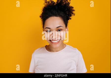 Nahaufnahme einer glücklichen afroamerikanischen jungen Frau mit lockigen Haaren und Sommersprossen, die ein einfaches T-Shirt trägt, auf einem isolierten orangefarbenen Hintergrund steht, direkt auf die Kamera blickt und freundlich lächelt Stockfoto