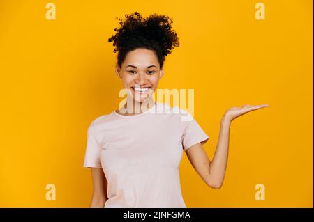 Foto einer freundlichen afroamerikanischen jungen Frau im lässigen T-Shirt, blickt auf die Kamera und zeigt die Hand zur Seite, auf leeren Raum für Präsentation oder Text, steht auf isoliertem blauen Hintergrund, lächelt Stockfoto