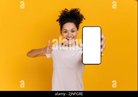 Verblüffte, lockige afroamerikanische junge Frau, in einem einfachen T-Shirt, zeigt ein Smartphone in der Hand mit einem weißen, weißen Mock-up-Bildschirm, steht auf einem isolierten orangefarbenen Hintergrund, blickt lächelnd auf die Kamera Stockfoto