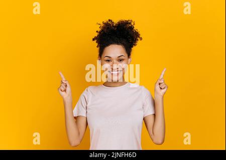 Positiv freundliche, afroamerikanische, lockige junge Frau, blickt auf die Kamera und zeigt die Finger nach oben, auf einen leeren Kopierraum, steht auf einem isolierten orangefarbenen Hintergrund und lächelt fröhlich Stockfoto