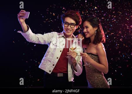 Liebevolles asiatisches Paar mit Sektflöten in den Händen, die Selfie auf dem Smartphone reden, während sie im Nachtclub ein bedeutsames Ereignis feiern, bunte Konfetti fliegen in der Luft Stockfoto