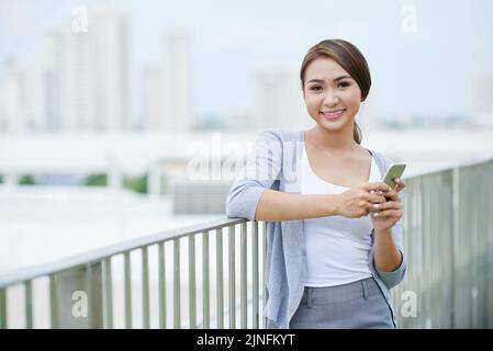 Porträt einer fröhlichen vietnamesischen jungen Frau mit lächelndem Smartphone und Blick auf die Kamera Stockfoto