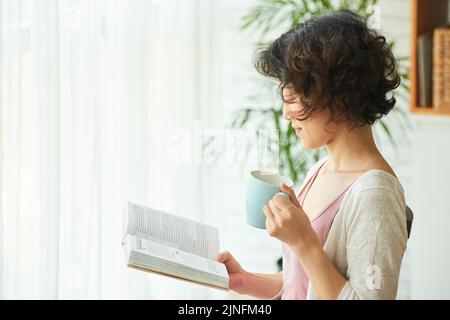 Profilansicht einer lockigen asiatischen Frau, die in eine Abenteuergeschichte eingehüllt wurde, während sie im gemütlichen Wohnzimmer mit einer Tasse Kaffee in der Hand saß Stockfoto