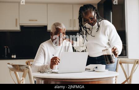 Unterstützende Frau, die ihrem Mann in seinem Heimbüro eine Tasse Kaffee ausgießt. Glückliche, reife Frau, die ihren Mann ermutigt, während er von zu Hause aus arbeitet. Stockfoto