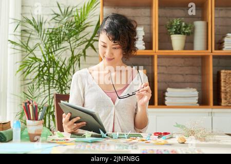 Attraktiver Designer mittleren Alters, der Dokumente studiert, während er am Schreibtisch eines modernen Designstudios sitzt, bunte Dekorelemente im Vordergrund Stockfoto
