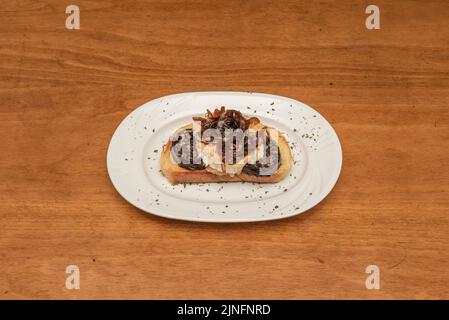 Black Pudding Toast mit Ziegenkäse und karamellisierten Zwiebeln, bestreut mit getrockneter Petersilie auf einem weißen Teller Stockfoto