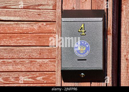 An der Wand montierter Briefkasten mit der Nummer 4, der an der roten Wand eines alten Holzhauses auf dem Land hängt. Die Inschrift auf Russisch, Post, für Briefe und Stockfoto